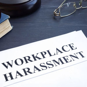 Paper with bold 'WORKPLACE HARASSMENT' text, glasses, book, gavel on dark surface. Urgent call to address this serious issue- Moss Bollinger LLP